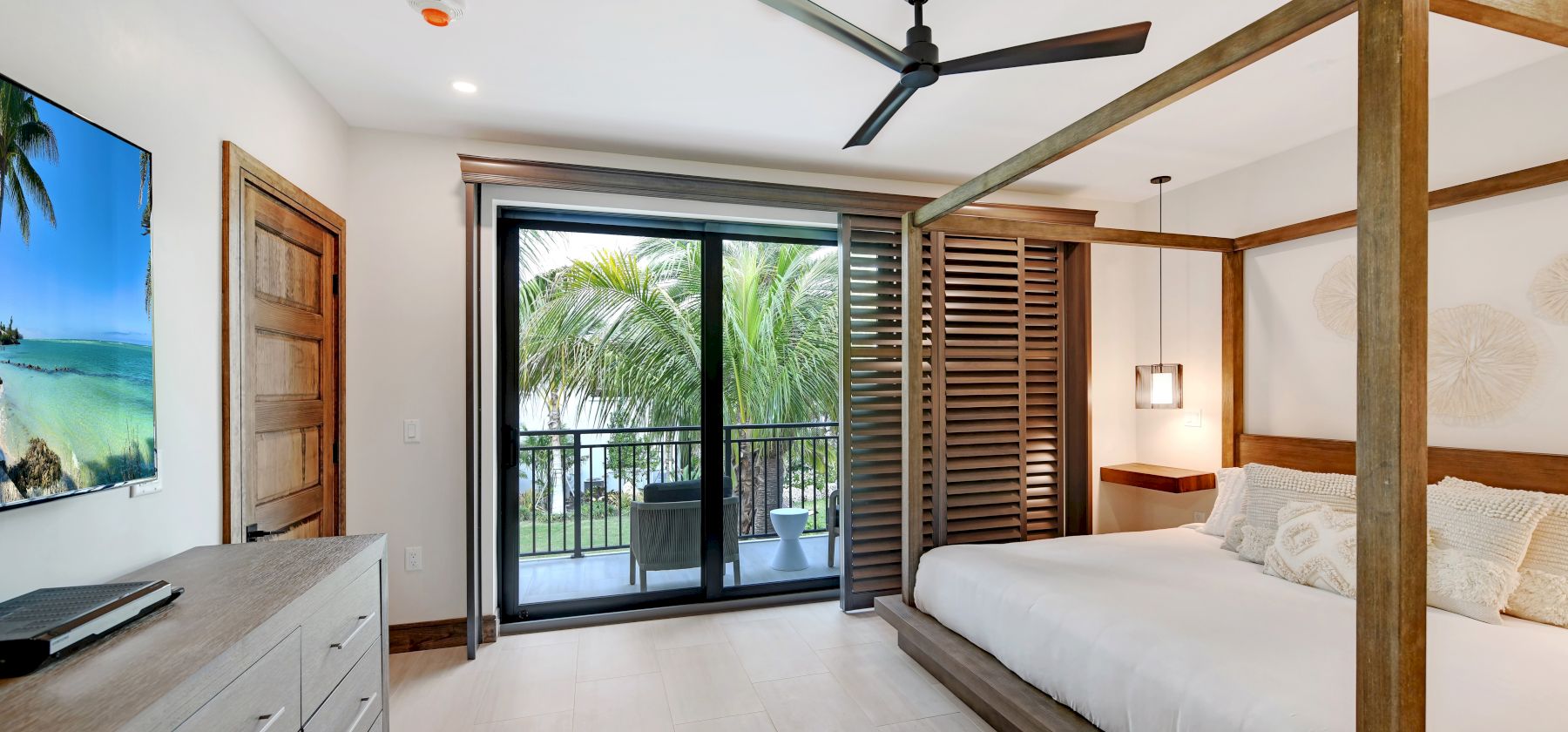 A bedroom with a wooden four-poster bed, ceiling fan, dresser with a TV, and glass doors leading to a balcony with a tropical view.
