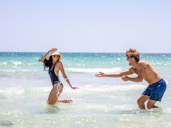 A man and woman are playfully splashing water on a sunny beach, with clear blue skies and waves in the background.