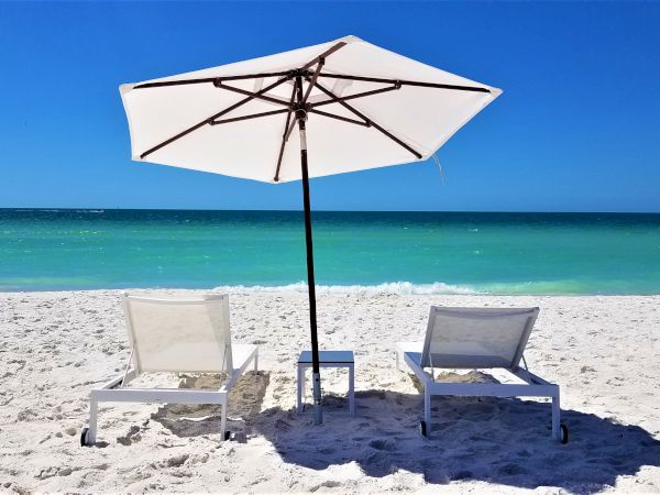 Two lounge chairs and a white umbrella on a sandy beach facing clear blue water under a bright sky.