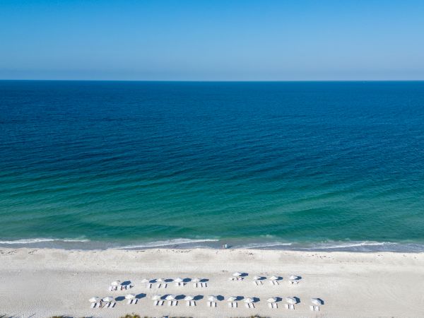 A tranquil beach with white sand, turquoise water near shore, and rows of blue-and-white umbrellas and lounge chairs facing the sea.