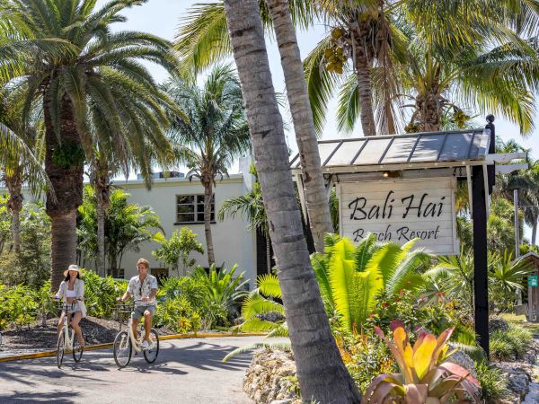 Two people ride bikes past a tropical resort entrance flanked by tall palm trees and lush plants, with a sign reading &ldquo;Bali Hai&rdquo; in the scene.
