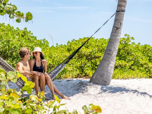 Two people sit in a hammock on a sunny beach, chatting and smiling under a leaning palm tree.