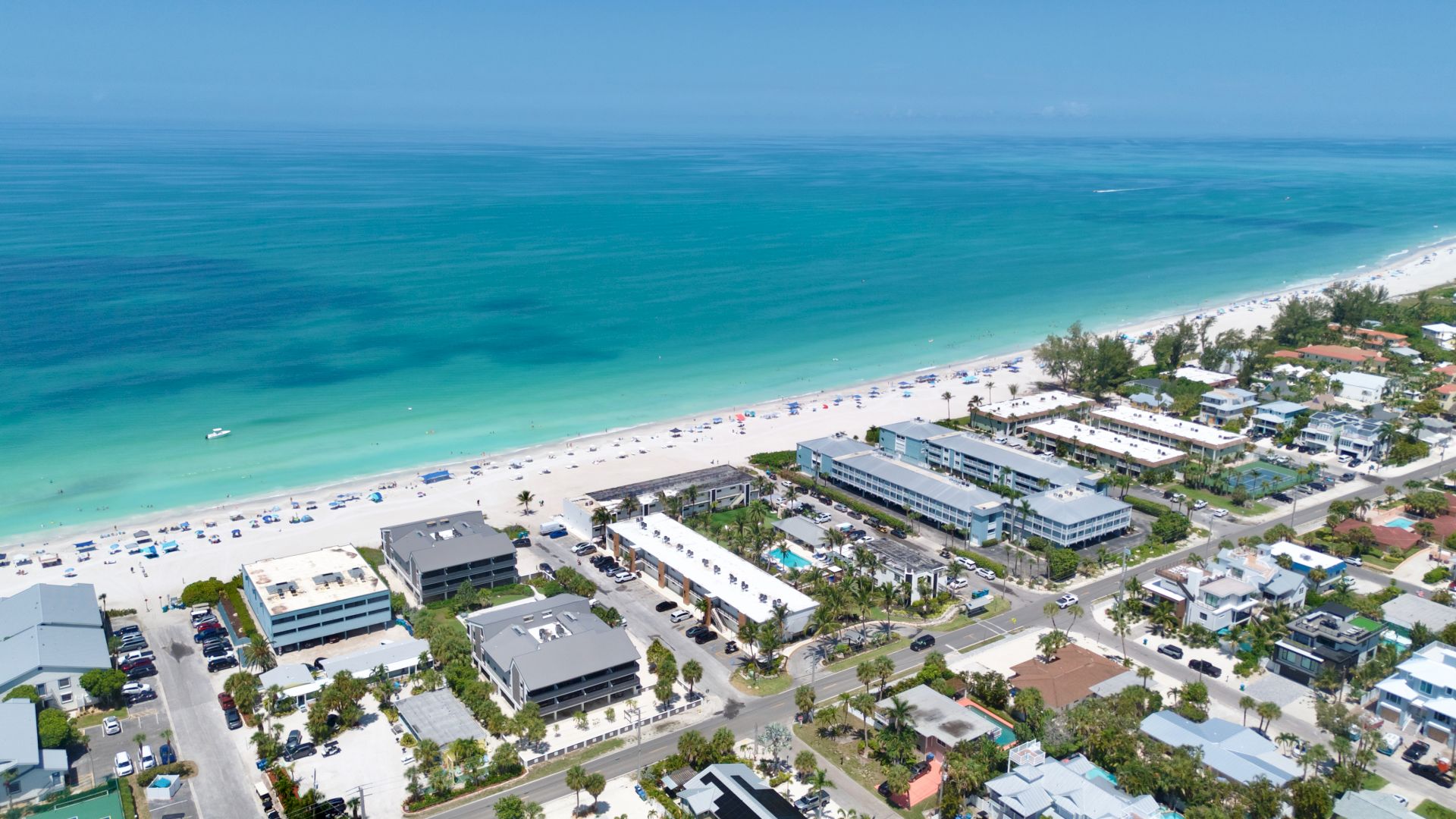 An aerial view of a tropical beach resort: turquoise ocean, sandy shoreline packed with umbrellas, and beachfront buildings.