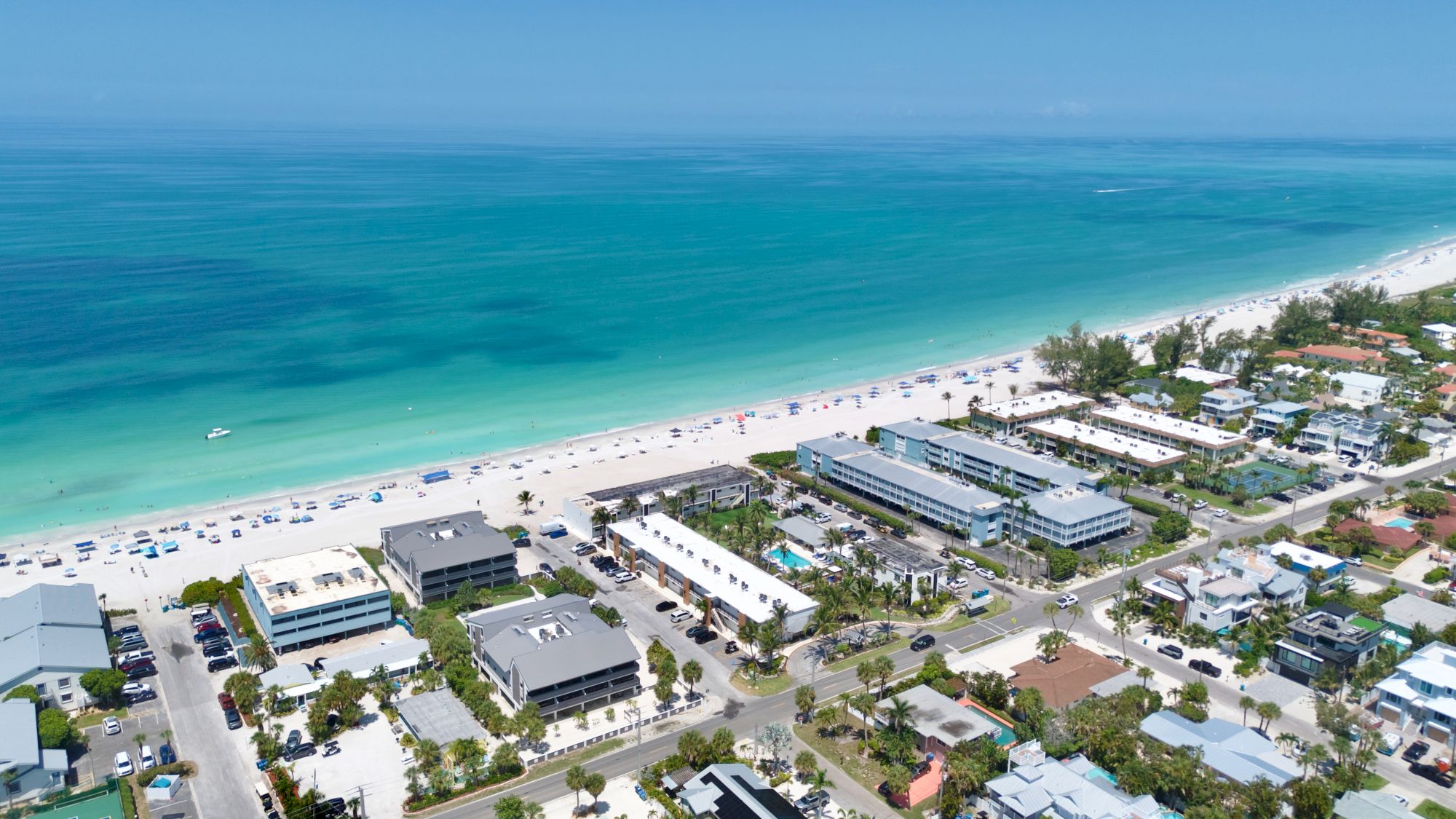 An aerial view of a tropical beach resort: turquoise ocean, sandy shoreline packed with umbrellas, and beachfront buildings.